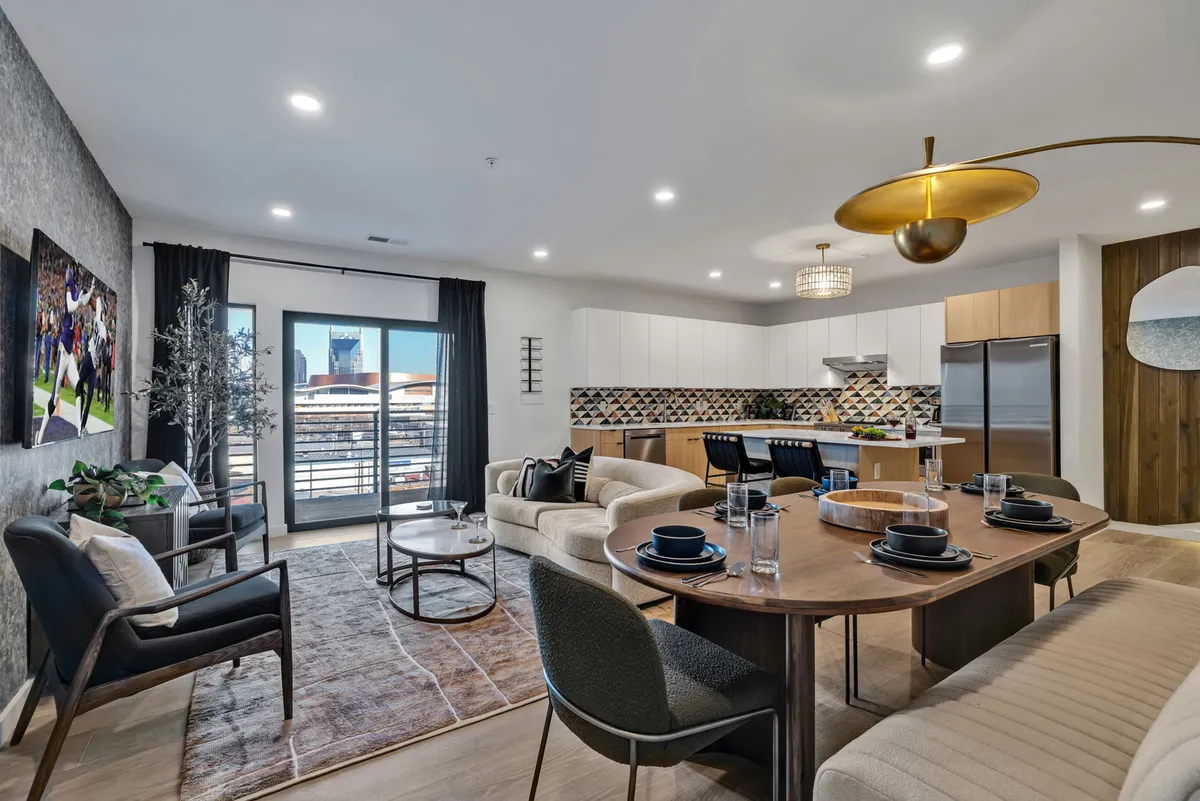 The image showcases a beautifully set dining table ready for a celebratory meal, with contemporary artwork adorning the wall, sleek modern kitchen appliances in view, and sliding glass doors leading out to a sunlit balcony. The space is open and inviting, perfect for gathering with friends or family.