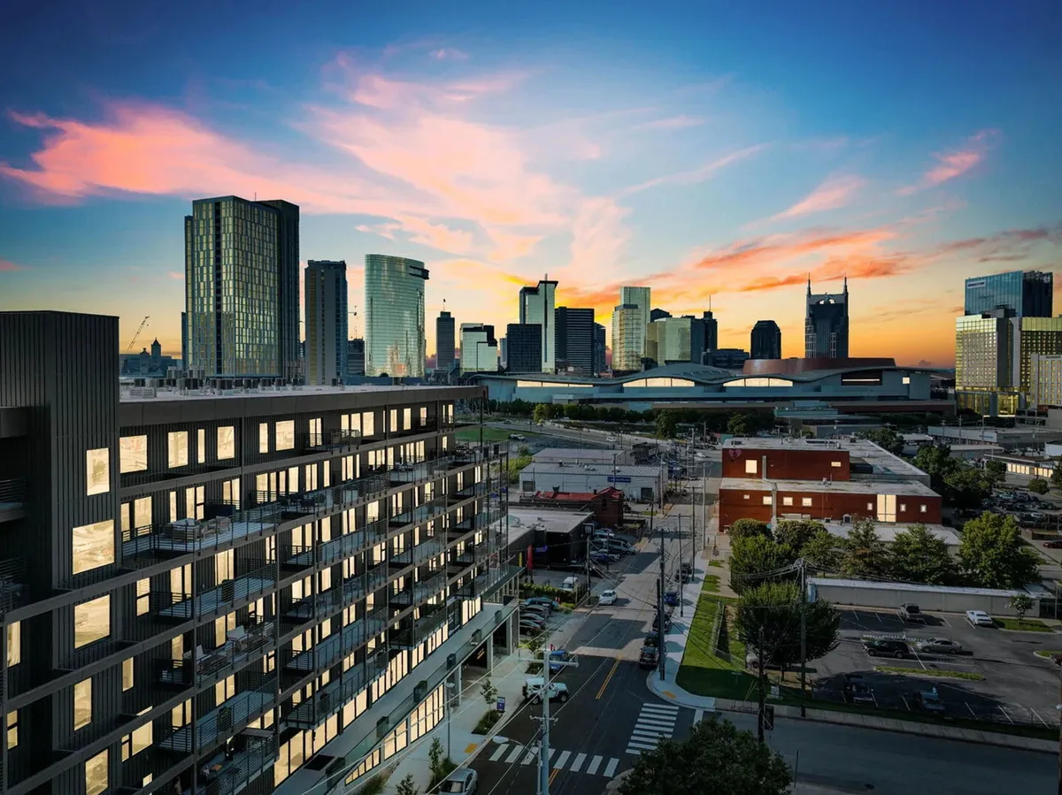 A striking view of a modern building against the Nashville skyline at sunset. The sky is filled with vibrant colors, and the city exudes an inviting energy as people enjoy evening strolls and outdoor dining in the area.
