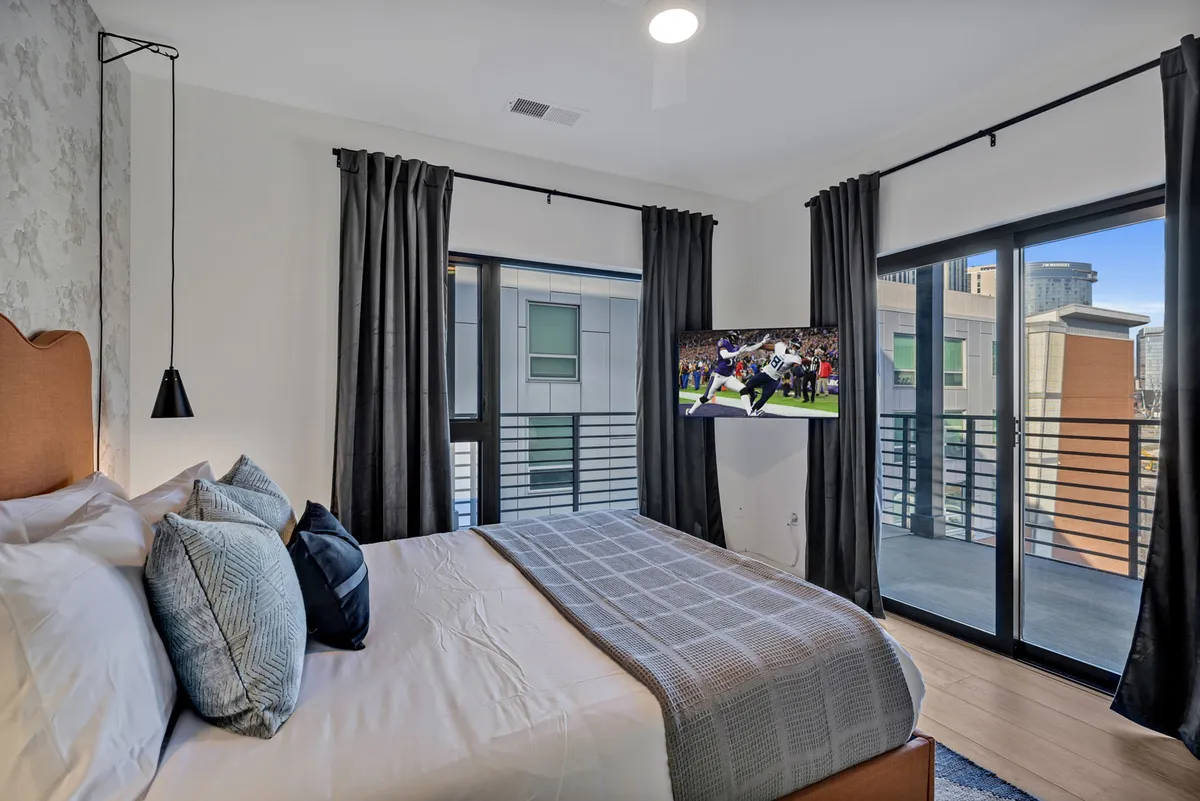 A bedroom featuring a comfortable bed with decorative pillows, a wall-mounted television, and large windows offering a view from the balcony. The setting is cozy and modern, hinting at private entertainment and access to outdoor space.