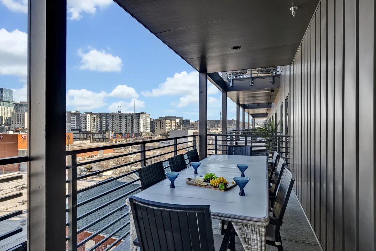 A bright, open balcony dining space with a table set with blue glasses and a view overlooking the Nashville skyline. The outdoor table is ready for gatherings, hinting at lively al fresco dining experiences and celebrations against a city backdrop.