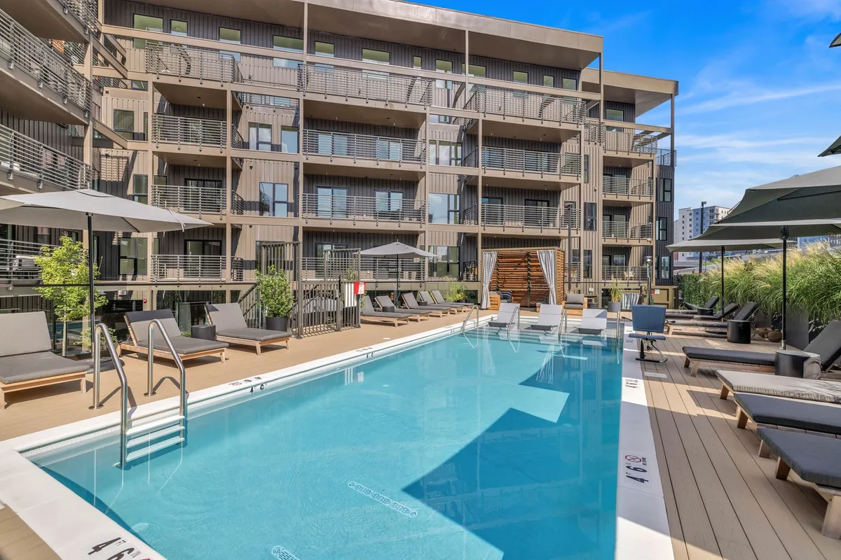 A vibrant exterior image of a swimming pool with lounge chairs, a building façade, and lush greenery. The scene is inviting and social, representing the pool and community grilling areas available at The Cosmo during pool season—a highlight for families looking to relax and have fun together.