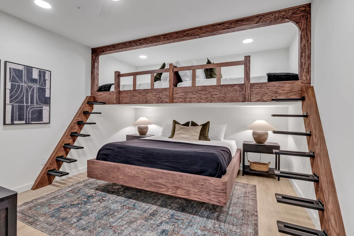 A playful and inviting bedroom with a lofted bed accessed by a wooden staircase, a soft textured rug, and cozy lighting from bedside lamps. The design encourages both relaxation and a sense of fun, suitable for families or groups.