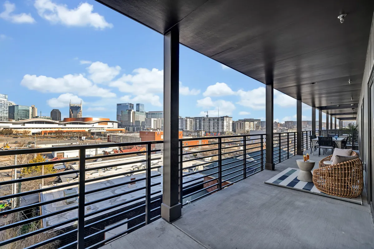 A private balcony with cozy outdoor furniture overlooks the vibrant Nashville city skyline, capturing both the energy of downtown and the tranquility of a personal outdoor retreat. The cityscape is bathed in natural light, suggesting the perfect setting for morning coffee or evening cocktails.
