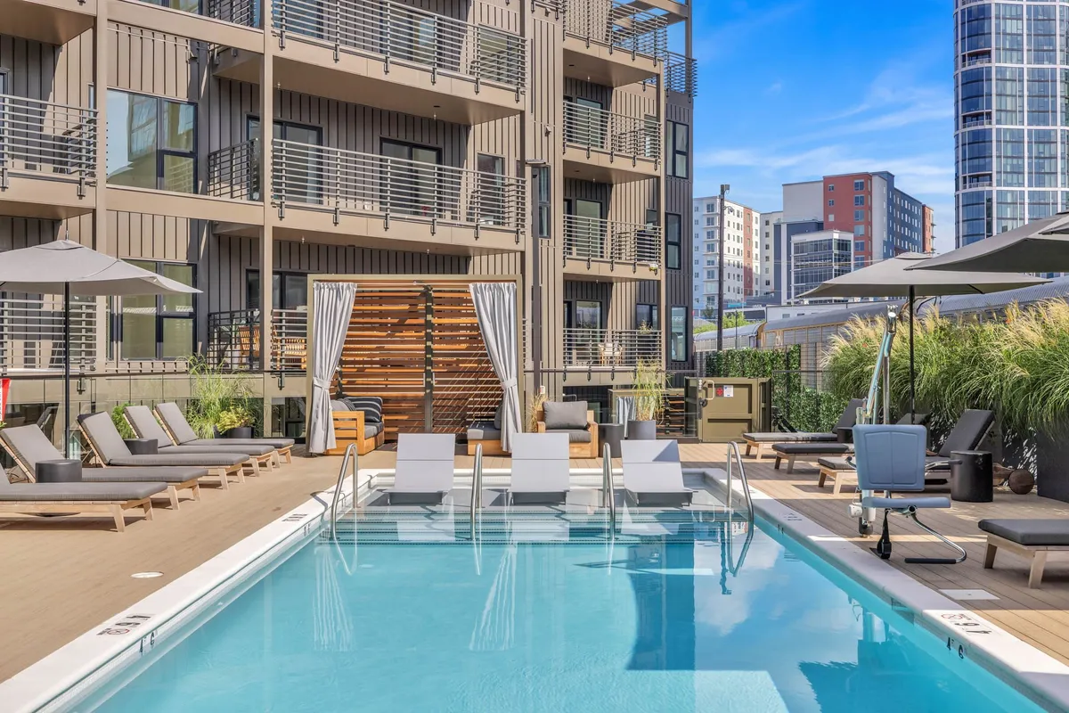 A vibrant outdoor pool area featuring inviting loungers, modern building facades, and comfortable seating spaces surrounded by greenery. The atmosphere is lively yet relaxed, perfect for sunny days and social gatherings.