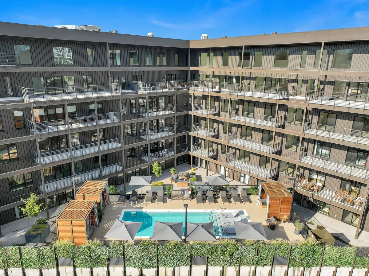 A sparkling outdoor swimming pool surrounded by lush greenery, modern lounge chairs, and comfortable seating areas, with hints of the building's facade in the background. The area is lively and designed for both daytime relaxation and social gatherings.