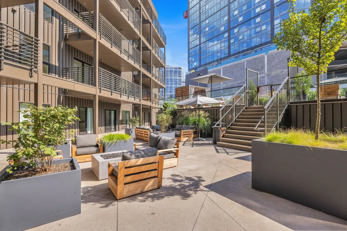 A contemporary outdoor seating area with cushioned benches, lush plants, and a welcoming atmosphere. The modern building facade and open space invite guests to relax, socialize, or enjoy an evening outdoors.