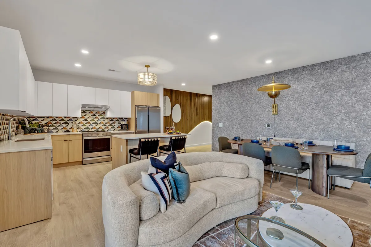 A contemporary setting with a curved sofa adorned with decorative pillows, a dining table stylishly set for a meal, and a kitchen area with a colorful backsplash and striking pendant lights. The space is bathed in natural light, creating a vibrant yet inviting atmosphere ideal for group gatherings.